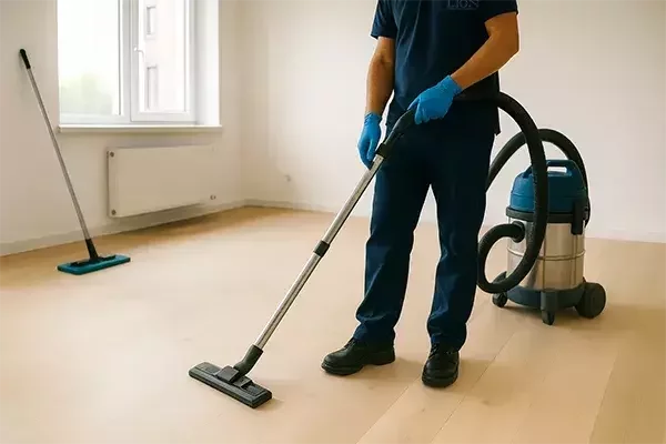 A newly built, unfurnished room with light wooden flooring being cleaned. A professional vacuum cleaner with hose attached and a mop are positioned near scattered dust on the floor, representing the process of after builders dust removal.