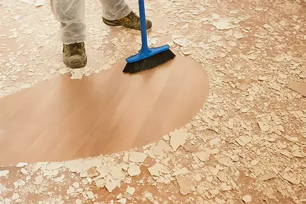 A cleaning technician sweeps up plaster debris with a blue broom, revealing a clean section of wooden floor during post-construction cleaning.