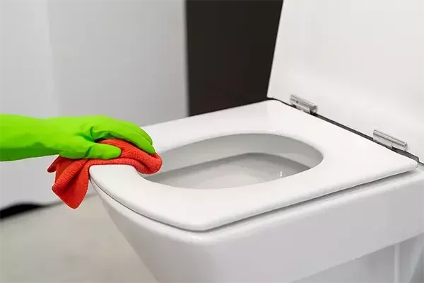 A person wearing a green glove wipes the rim of a white toilet with a red cleaning cloth.