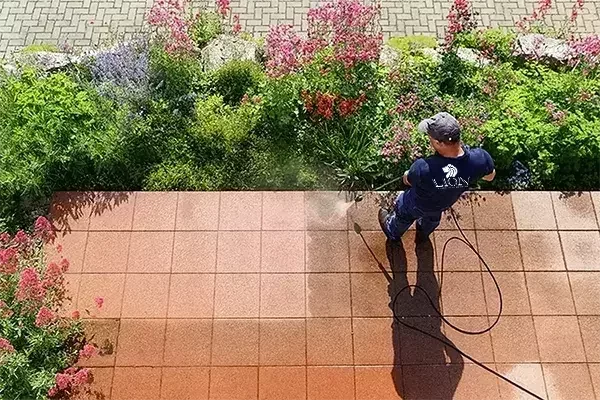 A person uses a pressure washer to clean outdoor patio tiles, with a clear contrast between the cleaned and uncleaned areas, next to a garden with colorful flowers and plants.