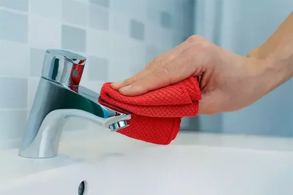 A hand wipes a chrome bathroom tap with a red cloth, leaving the surface polished and hygienic.