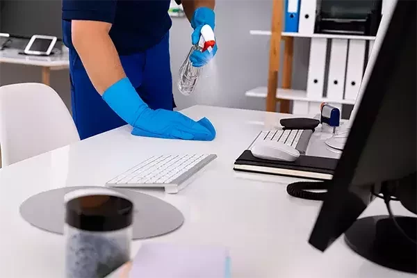 Cleaning operative wearing blue gloves sprays disinfectant and wipes down an office desk with a blue cloth, sanitising around a computer keyboard and workspace.
