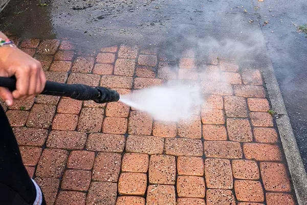 Cleaning technician uses a high-pressure washer to remove moss and dirt from outdoor brick paving, with water spray visibly lifting debris.