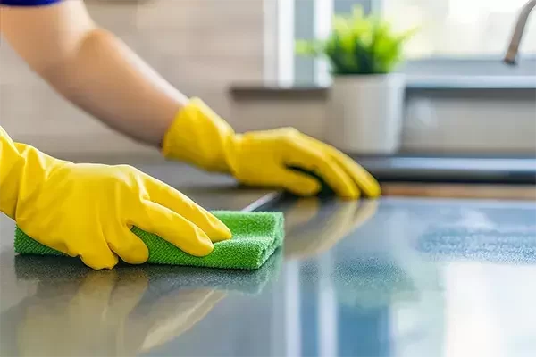 Hands wearing yellow gloves wiping a kitchen countertop with a green cloth to clean and disinfect the surface.