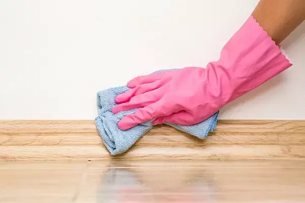 Hand wearing a pink rubber glove wiping a wooden skirting board with a blue cloth to remove dust and dirt.