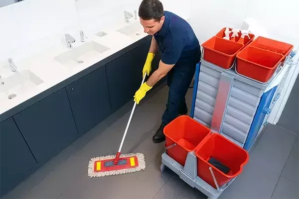 Cleaning operative wearing yellow gloves mops the tiled floor of a restroom, with sinks in the background and a cleaning trolley stocked with supplies beside them.