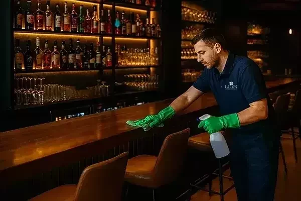 A cleaner wearing green gloves, wiping a wooden bar counter with a green cloth and spray bottle, with shelves of bottles and glasses in the background.