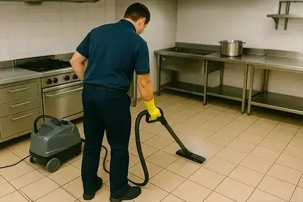 A cleaning operative, seen from behind, uses a commercial steam cleaning machine on the tiled floor of a stainless steel commercial kitchen.