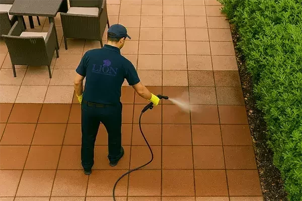 A cleaning technician in a navy uniform and yellow gloves pressure washes outdoor patio tiles, revealing a clear contrast between the cleaned and dirty areas beside a row of green bushes.
