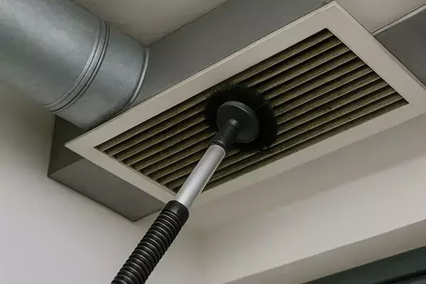 Rotary brush vacuum tool cleaning dust from an air vent grille connected to industrial ductwork.