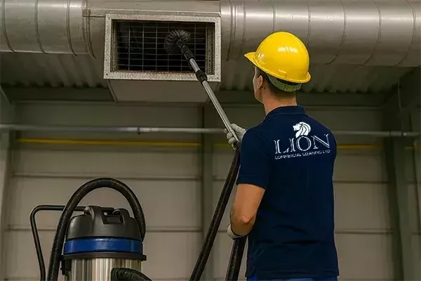 A cleaning technician in a yellow hard hat and high-visibility vest uses a vacuum with a brush attachment to clean an industrial air vent inside a facility.