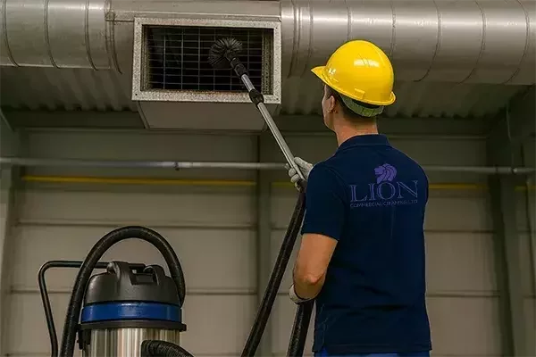 A cleaning technician in a yellow hard hat and high-visibility vest uses a vacuum with a brush attachment to clean an industrial air vent inside a facility.
