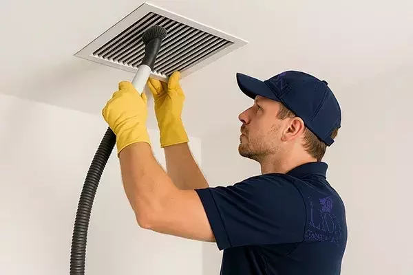 A cleaning technician wearing a navy shirt, cap, and yellow gloves uses a vacuum hose with a brush attachment to remove dust from a ceiling air vent.