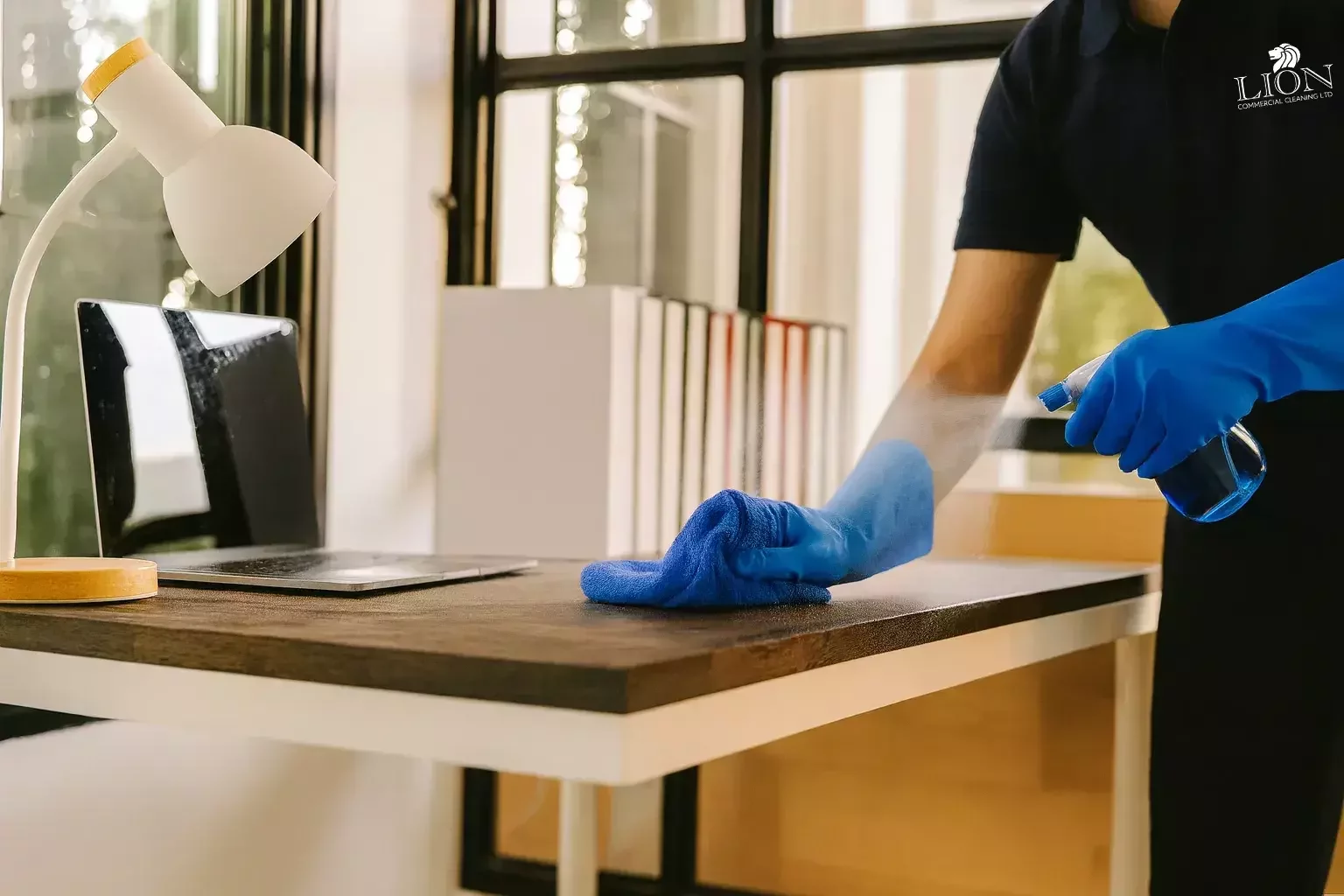 A cleaning technician wearing blue gloves sprays disinfectant and wipes down an office desk with a cloth, next to a laptop, desk lamp, and books, ensuring a hygienic workspace.