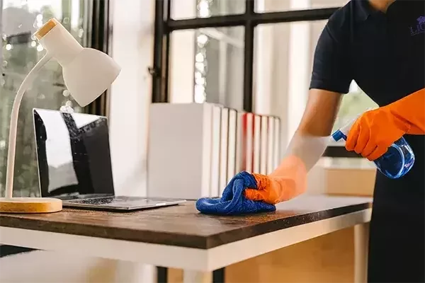 A cleaning technician wearing orange gloves sprays disinfectant and wipes down an office desk with a cloth, next to a laptop, desk lamp, and books, ensuring a hygienic workspace.