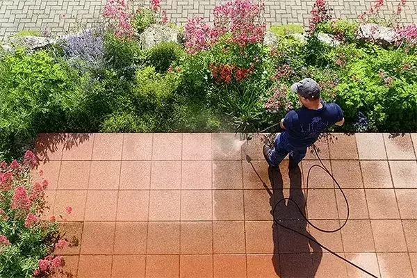 A cleaning operative uses a pressure washer to clean outdoor tiles, revealing a clear contrast between the cleaned and dirty sections, with a garden full of colorful flowers in the background.