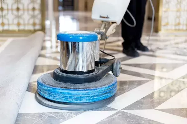 A professional floor polishing machine with a blue pad being used on a patterned marble floor, leaving the surface shiny and clean as a rolled-up carpet sits to the side.