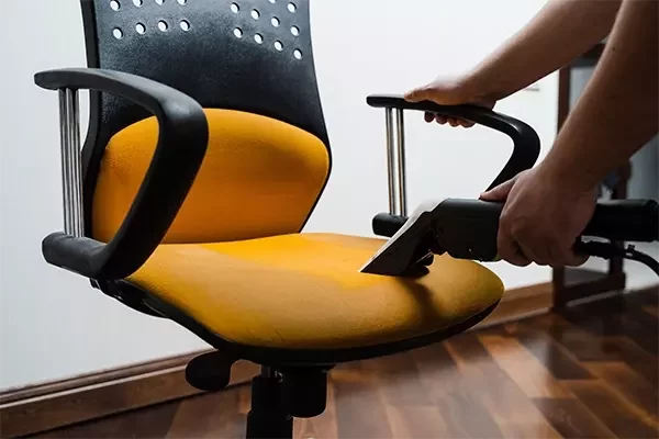 A cleaning technician uses a professional extraction tool to deep clean the fabric seat of a yellow office chair, removing dirt and stains to restore its appearance.