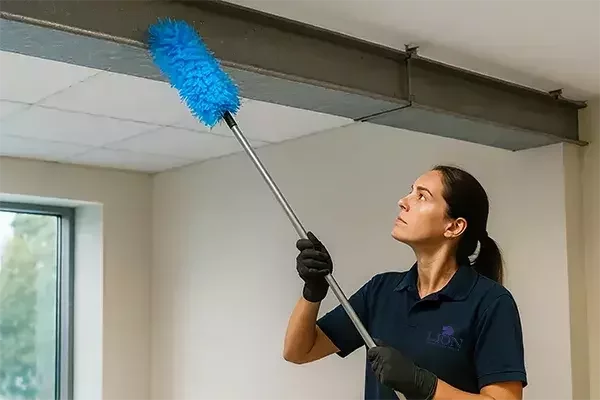 An office cleaning specialist wearing a navy shirt and black gloves uses a long blue duster to remove dust from high ceiling beams in an office space.