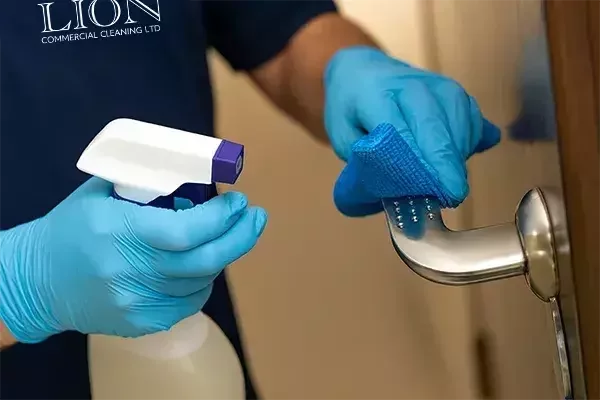 A cleaning operative wearing blue gloves disinfects a stainless steel door handle with a spray bottle and blue cloth, ensuring hygiene on a high-touch surface.