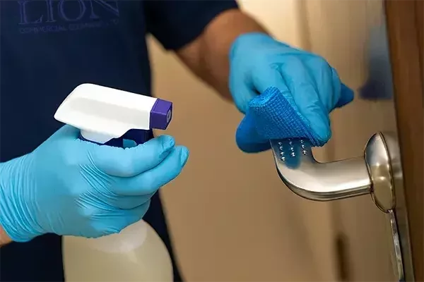 A cleaning operative wearing blue gloves disinfects a stainless steel door handle with a spray bottle and blue cloth, ensuring hygiene on a high-touch surface.