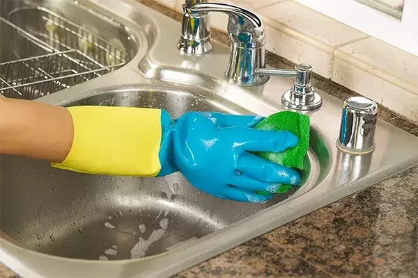A cleaning operative wearing yellow and blue rubber gloves scrubs a stainless steel kitchen sink with a soapy sponge, focusing on hygiene and shine.