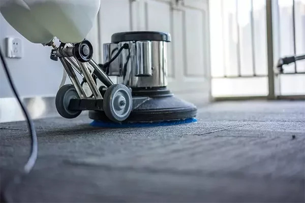 A close-up of a professional floor cleaning machine scrubbing a carpeted surface, with visible soap and water being worked into the fibers for deep cleaning.