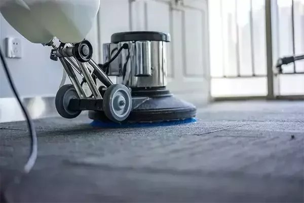 A close-up of a professional floor cleaning machine scrubbing a carpeted surface, with visible soap and water being worked into the fibers for deep cleaning.