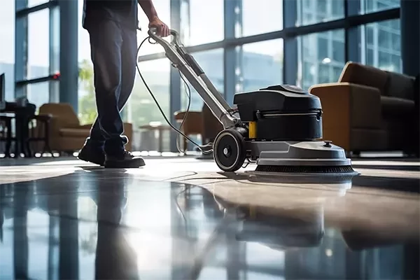 A janitorial worker operating a professional floor polishing machine on a glossy tiled surface in a modern office or lobby, with large windows and seating visible in the background.