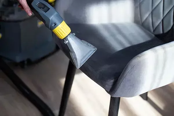 A cleaning technician using a professional extraction tool to deep clean the fabric of a grey upholstered chair, removing dirt and stains from the seat.