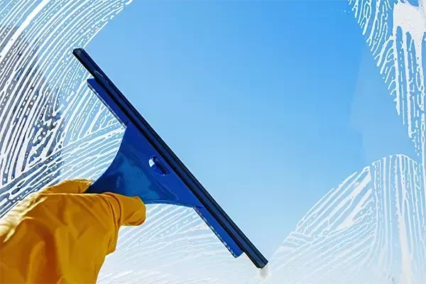 A janitorial worker wearing yellow gloves uses a blue squeegee to remove soap and water from a glass window, leaving a clear, streak-free finish against a bright blue sky.
